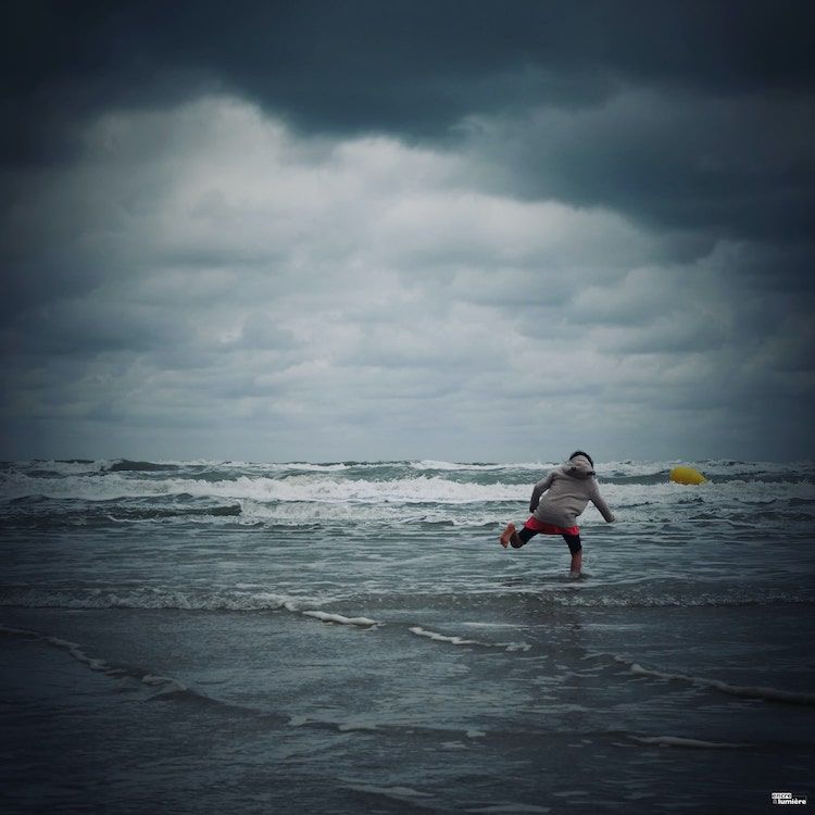 Photo d'un enfant jouant dans les vagues, temps gris, nuages. Photographe normandie Antoine Fantin.