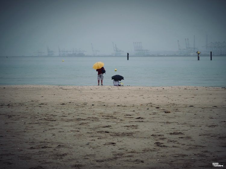 Couple avec des parapluies sous la pluie sur la plage de Honfleur. On aperçoit le port du Havre dans les brumes.Série "Un temps de chien" de Antoine Fantin.